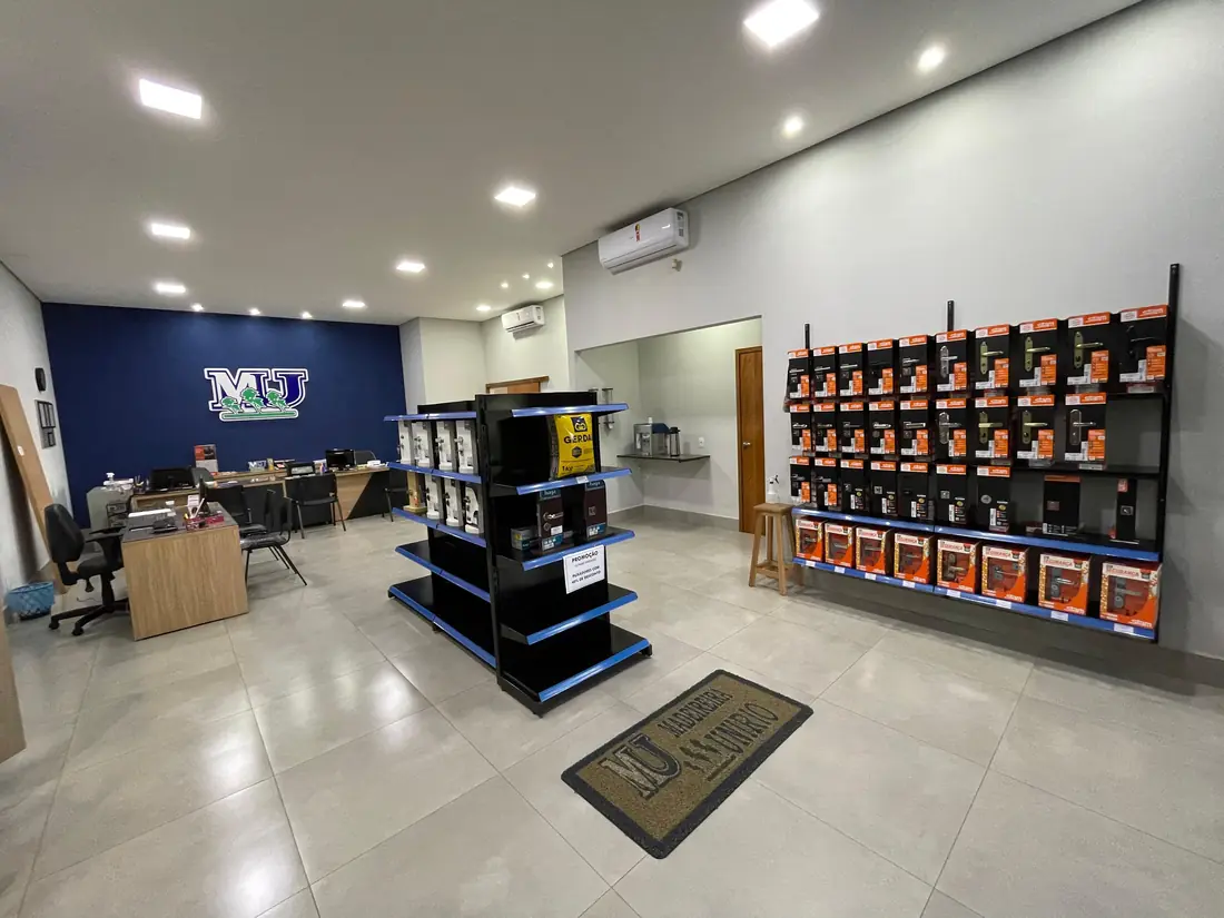 Salão de atendimento da Madeireira Unirio: piso claro, parede azul com o monograma MU, gôndola central de prateleiras azuis e uma parede lateral com ferragens expostas em embalagens.